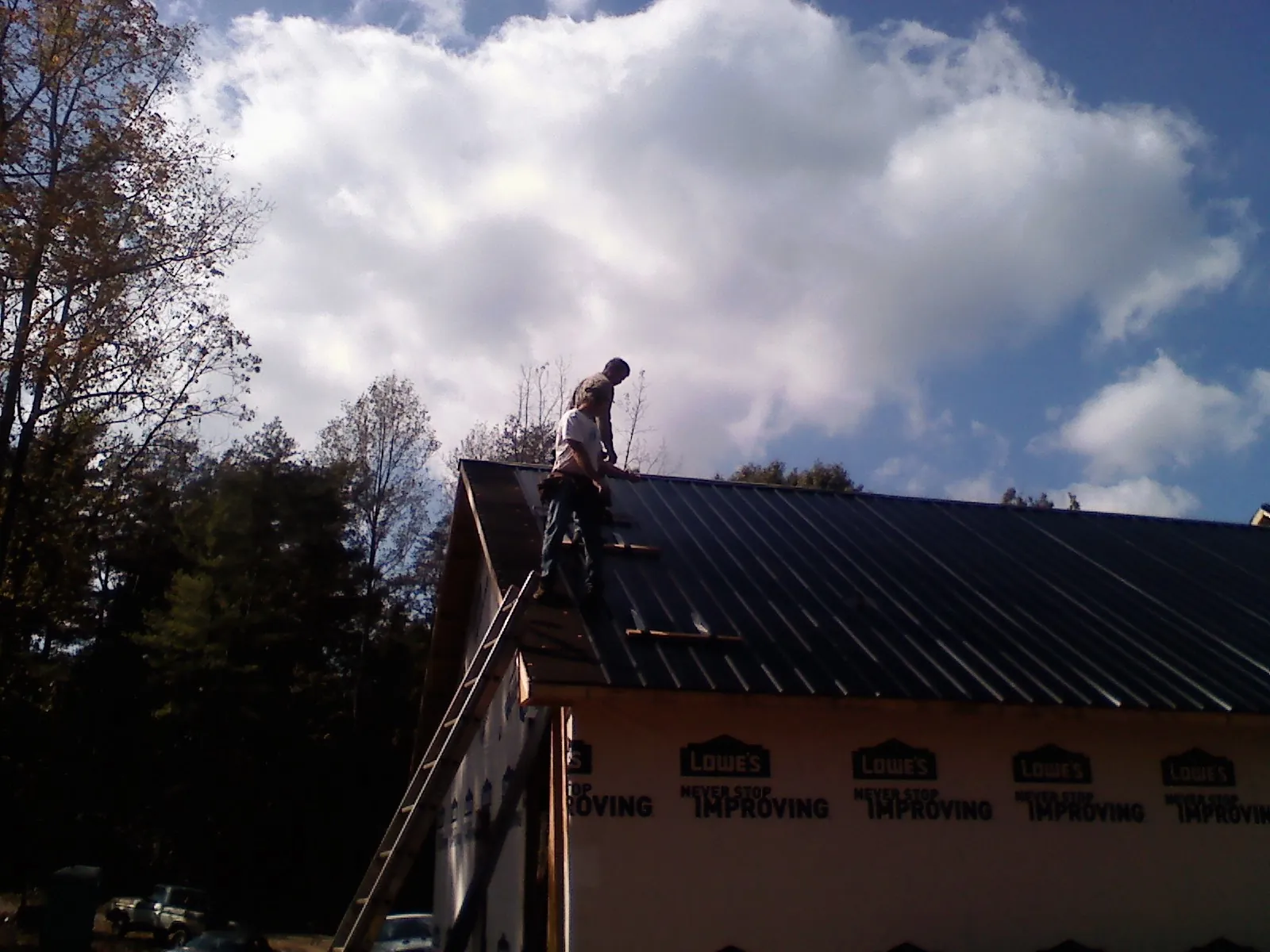 Roofer fastening dark metal panels high against a cloudy sky, from a Roof Inspection job in Olney